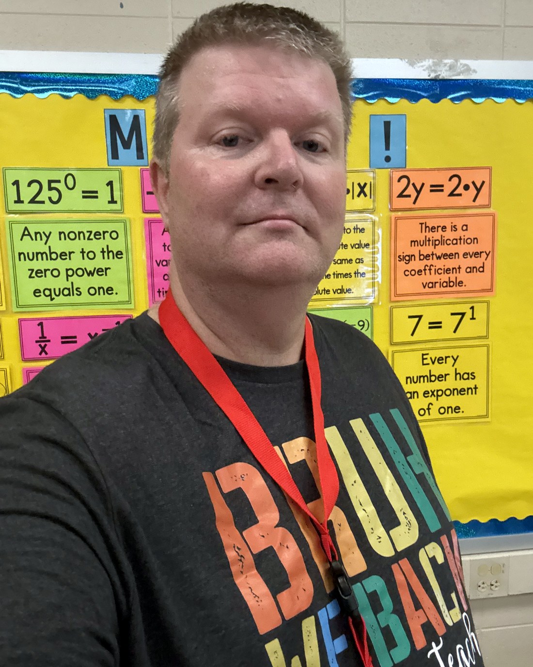 Greg, the founder of Math Class 678, in his middle school math classroom in front of anchor charts for exponent rules and the distributive property